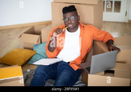 Pensive Black Young Man Sitting Among Cardboard Boxes And Using Laptop Stock Photo