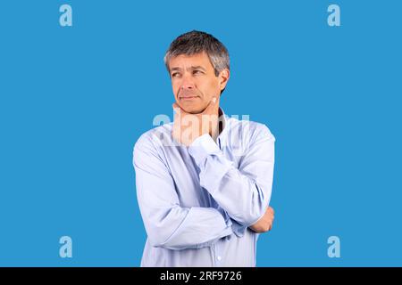 Pensive mature man touching chin with thoughtful expression, blue background Stock Photo