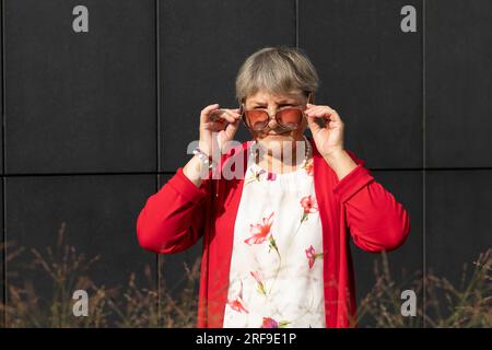 Real Cute Senior Old 70s Woman Looking Over Sunglasses Outdoor In Summertime, Dark Wall on Background. Portrait Of Smiling Kind Elderly Female Stock Photo