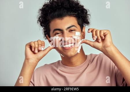 Studio portrait of happy smiling guy with black curly hair applying cream on his face. Handsome biracial swarthy man uses beauty products in his skinc Stock Photo