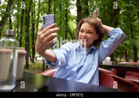 Attractive smiling mature woman making selfie or video call. Senior lady uses smartphone. Middle aged brunette female in blue shirt at the table in ou Stock Photo