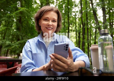 Attractive smiling mature woman making selfie or video call. Senior lady uses smartphone. Middle aged brunette female in blue shirt at the table in ou Stock Photo