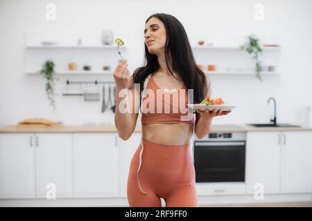 Portrait of slim beautiful fitness model posing with plate of nutritious snack and smiling at camera. Good-looking woman in tight sport clothing and measuring tape on neck leading healthy lifestyle. Stock Photo