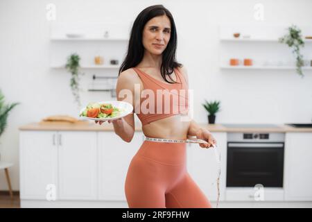 Charming lady controlling weight and measuring waist with tape after finishing morning workout. Content woman in tight sport clothes holding plate with nutritious meal and rejoicing amazing results. Stock Photo
