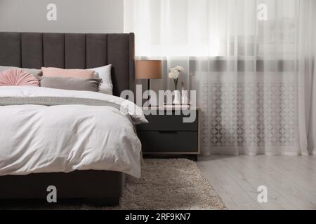Comfortable bed with cushions, lamp and different decor on bedside table in room. Stylish interior Stock Photo
