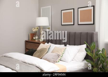 Comfortable bed with cushions, lamp and different decor on wooden chest of drawers in room. Stylish interior Stock Photo