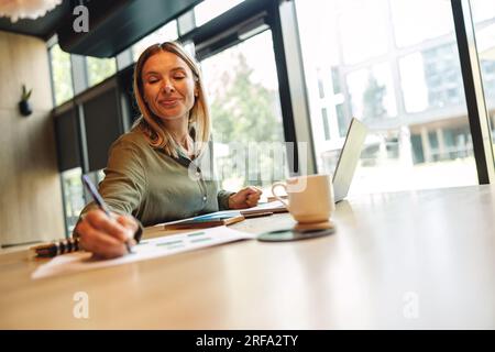 Pretty woman entrepreneur making notes in note pad while sitting in cozy coworking Stock Photo
