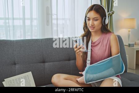 Young asian woman wearing soft splint due to sore arm using phone and listening music on sofa at home. Stock Photo