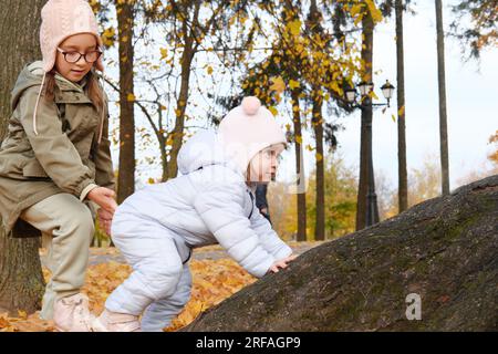 Two sisters are trying to climb a tree in the autumn park. The older sister helps the younger. Horizontal photo Stock Photo