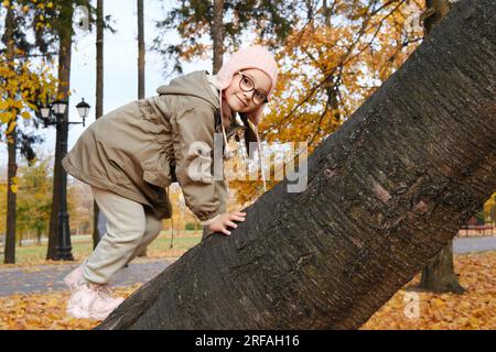 Girl child in glasses, a pink hat and an autumn jacket climbs up a tree in an autumn park. Horizontal photo Stock Photo