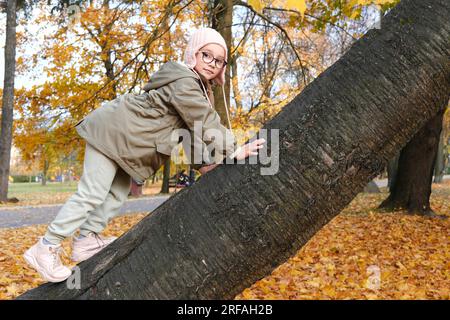 Girl child in glasses, a pink hat and an autumn jacket climbs up a tree in an autumn park. Horizontal photo Stock Photo