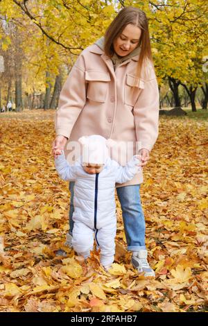 Mother spends time with her daughter in the autumn park. Mother supports her daughter by the hands and helps her to walk. Vertical photo Stock Photo