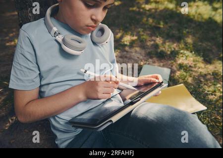 Handsome smart school boy using a digital tablet and stylus pen, drawing, sketching, enjoying his creative hobby outdoors. People. Leisure activity. L Stock Photo