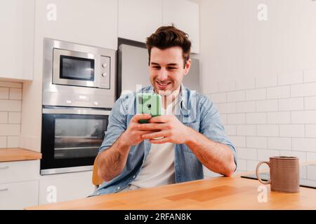 Happy caucasian guy using his smart phone chatting sending messages on social media sitting at home kitchen and taking a break. Smiling young man playing a video game on cellphone or texting an email. High quality photo Stock Photo
