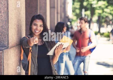 Beautiful latin american female student with digital tablet computer and other young adults outdoor in front of university building Stock Photo