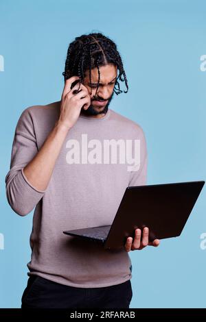 Puzzled arab programmer solving code problem on laptop and rubbing head. Young adult person with unsure facial expression holding portable computer and developing software Stock Photo