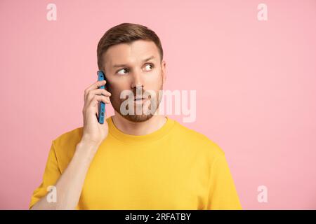Puzzled frustrated man in yellow t-shirt talking on phone on pink background with empty space Stock Photo