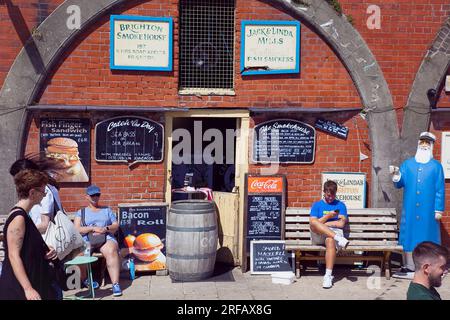 England, East Sussex, Brighton, Smokehouse fish sandwich vendor in arches by seafront promenade busy with summer tourists. Stock Photo