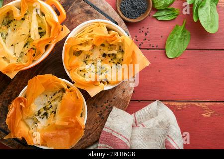 Filo pies with soft feta cheese and spinach in ceramic molds on old red wooden table backgrounds. Filo portions pies. Small Baked Spanakopita pies. To Stock Photo