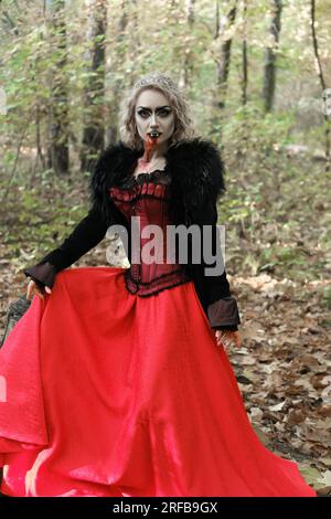 A woman in the image of a vampire or a witch with white make-up and in a medieval dress poses in the forest in front of the camera. Vertical photo Stock Photo