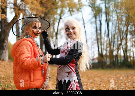 A girl dressed as a cheerleader and with half-face makeup in the form of a skeleton adjusts the hat of a girl in the form of a pumpkin. Girls smiling Stock Photo