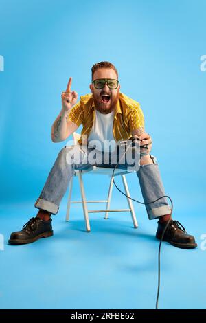 Bearded man in casual clothes and sunglasses emotionally playing console against blue studio background. Evening fun Stock Photo
