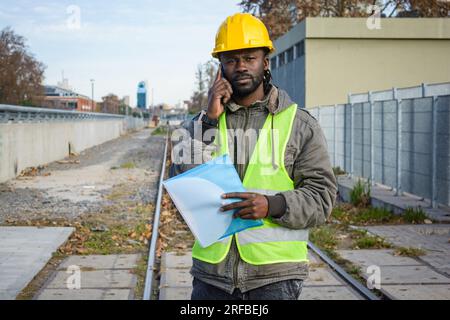 portrait of black man maintenance worker wearing helmet and reflective vest on a call talking on the phone seriously standing on the railway tracks, c Stock Photo