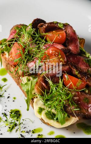 Bruschetta with juicy meat, tomatoes, herbs, cheese, pesto sauce on a white plate. Close-up. Stock Photo