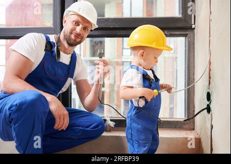 Male electrician holding electrical plug and smiling while child using electric soldering iron tool. Man and kid wearing work overalls and safety helmets while working on home renovation. Stock Photo