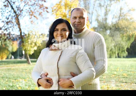 Husband and wife in white warm clothes pose for the camera in an autumn park on a sunny day. Husband hugging his wife from behind. Horizontal photo Stock Photo