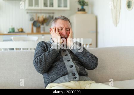 Headache pain. Unhappy middle aged senior man suffering from headache sick rubbing temples at home. Mature old senior grandfather touching temples experiencing stress. Man feeling pain hurt in head Stock Photo