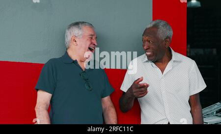 Joyful Interaction of Two Diverse Male Senior Friends, Smiling and Laughing Together, Leaning on Sidewalk Wall. Authentic real life happy elderly peop Stock Photo