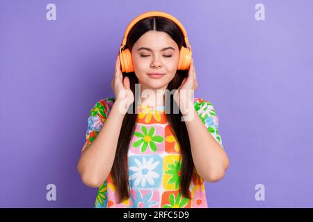 Photo of satisfied girl straight hairdo dressed flower print t-shirt touching headphones enjoy music isolated on violet color background Stock Photo