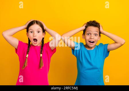 Photo of impressed shocked preteen schoolkids dressed t-shirts arms head open mouth isolated yellow color background Stock Photo