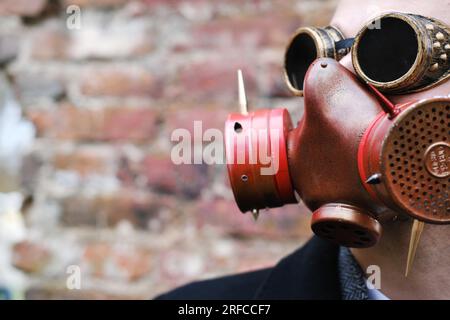 A man in a black coat, business suit, gas mask and steampunk goggles poses against a brick wall. Horizontal photo Stock Photo
