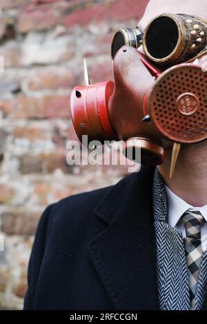 A man in a black coat, business suit, gas mask and steampunk goggles poses against a brick wall. Vertical photo Stock Photo