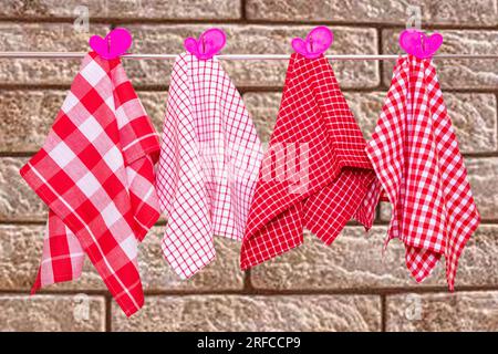 Hanging towels. Closeup of various red checkered kitchen towels with violet pink clothespins hang on a clothes rail against abstract blurred brick wal Stock Photo