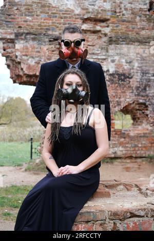 A young man in steampunk goggles, a gas mask and a woman with dreadlocks and a gas mask pose against the background of a destroyed brick wall. Vertica Stock Photo