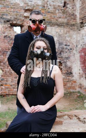 A young man in steampunk goggles, a gas mask and a woman with dreadlocks and a gas mask pose against the background of a destroyed brick wall. Vertica Stock Photo
