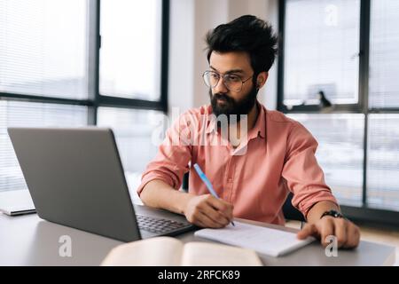 Successful bearded Indian businessman in glasses using laptop analyzing working project results, writing notes siting at table by window. Stock Photo