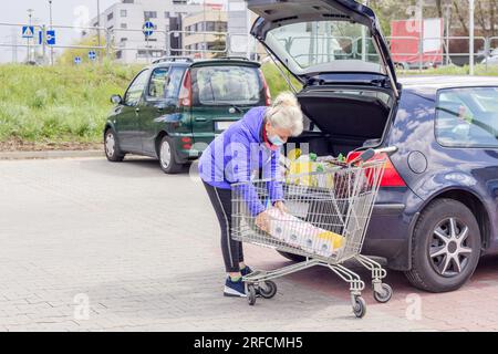 A mature good looking woman wearing protective mask loads toilet paper into a car in the parking lot. Everyday life during coronavirus pandemic. Stock Photo