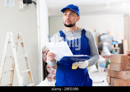 Responsible focused bearded supervisor wearing blue overall taking notes on papers while inspecting construction site Stock Photo