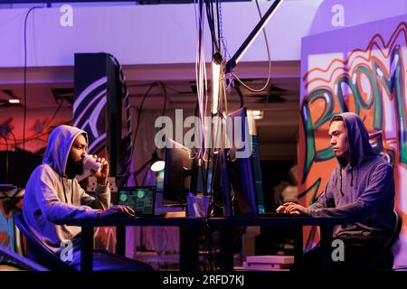 Hackers breaking law and doing illegal activity on computer late at night. Two young diverse criminals drinking coffee while programming malicious software in dark hideout place Stock Photo