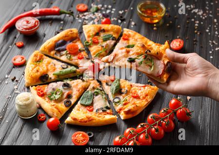 Hands take slices of cut pizza on a dark wooden background top view. Pizza with salami, jerky, sauces, spices, fresh cherry tomatoes on a black Stock Photo