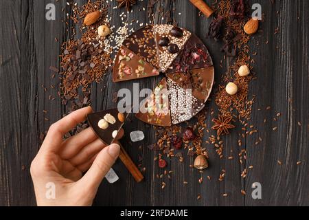 Chocolate pizza with nuts, dry berries, sesame seeds and ingredients on a dark wooden background top view. The hand holds a piece of sweet chocolate p Stock Photo