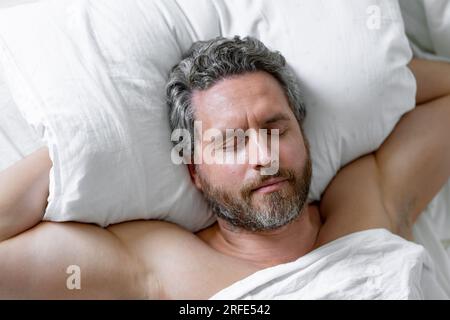 Male sleeps on pillow. Gray hair man sleep in white bed. Senior man sleep in bed at bedroom. Mature man morning sleeps at home. Good sleep. Millennial Stock Photo