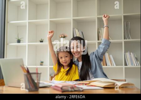 A portrait of a cheerful Asian female teacher and a happy little girl are raising hands together at a table in the classroom. positive classroom, havi Stock Photo