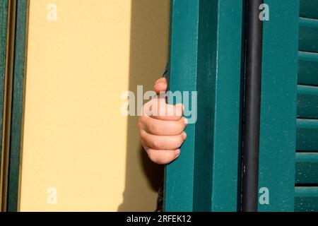 Closeup of child's hand holding on to petrol colored antique wooden door shutters in natural sunlight with yellow wall in the background Stock Photo