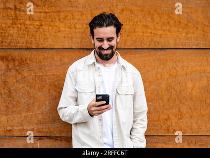 Confident handsome young guy leaning against a wooden wall while texting with his phone. Happy and relaxed man using his smartphone standing on a Stock Photo