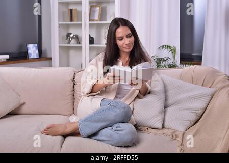 Woman engrossed in book on cozy living room sofa. Soft ambiance and peaceful expression reveal complete immersion in captivating world of literature Stock Photo